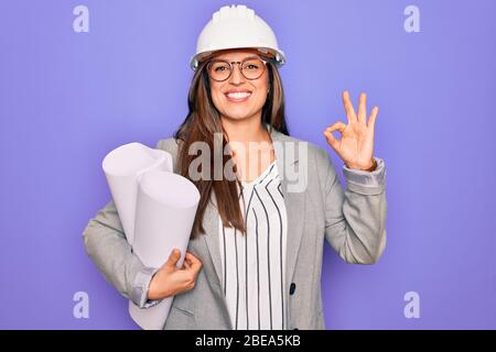 Junge hispanische Architektin Frau trägt Baumeister Hut und hält Blaupausen über gelbem Hintergrund tun ok Zeichen mit Fingern, ausgezeichnetes Symbol Stockfoto
