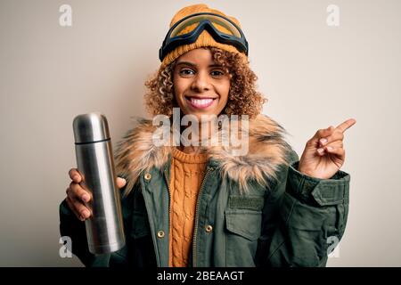 Junge afroamerikanische Skifahrer Frau mit Skibrille trinken Thermo mit Kaffee sehr glücklich mit Hand und Finger zur Seite zeigen Stockfoto