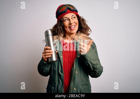 Frau mittleren Alters, die Skierbrille trägt, trinkt Thermo mit Kaffee auf weißem Hintergrund und zeigt mit dem Daumen bis zur Seite mit glücklicher Fac Stockfoto
