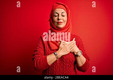 Frau mittleren Alters trägt traditionelle muslimische Hijab stehend über isolierten roten Hintergrund lächelnd mit Händen auf der Brust mit geschlossenen Augen und dankbar gest Stockfoto