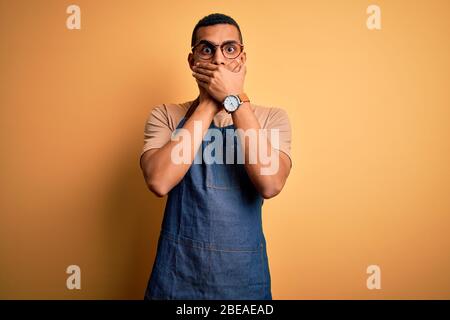 Junge gut aussehende afroamerikanischen Ladenbesitzer Mann trägt Schürze über gelben Hintergrund schockiert Abdeckung Mund mit den Händen für Fehler. Geheimkonzept. Stockfoto