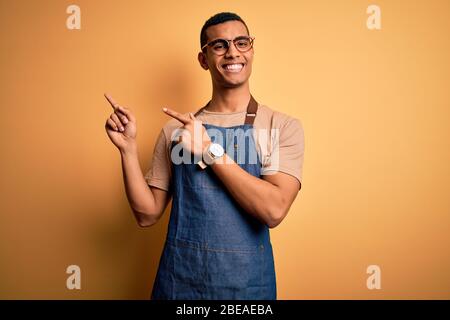 Junger schöner afroamerikanischer Ladenbesitzer, der Schürze über gelbem Hintergrund trägt und lächelnd auf die Kamera schaut, mit zwei Händen zeigt und fing Stockfoto