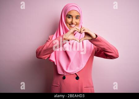Junge schöne Brünette Geschäftsfrau trägt rosa muslimischen Hijab und Business-Jacke lächelnd in Liebe tun Herz Symbol Form mit Händen. Romantischer Nachteil Stockfoto