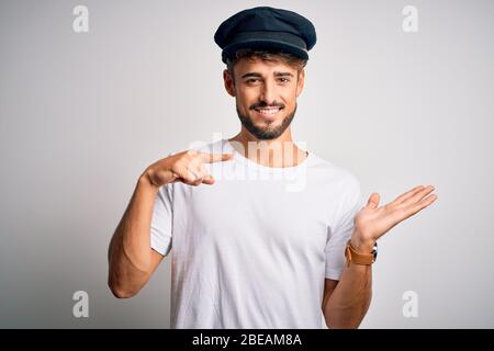 Junger Fahrer Mann mit Bart trägt Hut über isoliert weißem Hintergrund erstaunt und lächelnd auf die Kamera, während mit Hand und Pointi präsentieren Stockfoto