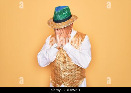 Mittelalter Senior grauhaarige Mann trägt brasilianischen Karneval custome über gelben Hintergrund mit traurigen Ausdruck bedeckt Gesicht mit Händen während weinen. Stockfoto