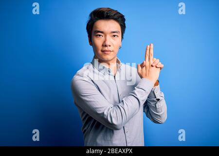 Junge gut aussehende chinesische Mann mit lässigem Hemd über isolierten blauen Hintergrund Halten symbolische Waffe mit Handbewegung, Töten Schießerei spielen Stockfoto