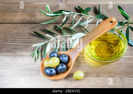 Grüne und schwarze Oliven in Holzlöffel, Olivenöl Glasschüssel und Olivenbaum Zweige auf dem Holzhintergrund. Schöne leichte, gesunde Ernährung. Stockfoto