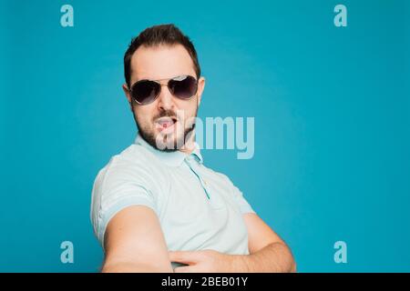 Ein junger bärtiger Mann täuscht sich auf blauem Hintergrund vor der Kamera herum. Der Kerl nimmt ein selfie und baut lustige Gesichter. Stockfoto