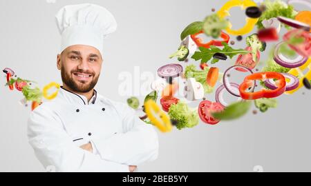 Gesunde Rezepte. Fröhlicher Koch trägt weiße Uniform und Gemüse in der Luft auf grauem Hintergrund. Collage Stockfoto