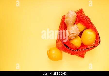 Frische Zitronen und Ingwerwurzel in einem roten Beutel zum Verpacken von Gemüse und Obst auf gelbem Hintergrund. Selektiver Fokus. Stockfoto