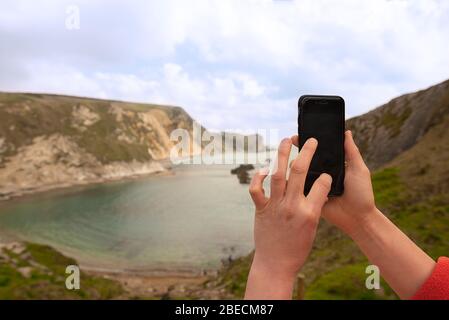 Weibliche Hände machen ein Foto auf einem Smartphone von man O'war Beach neben Durdle Door an der Jurassic Coast, Südengland. Stockfoto