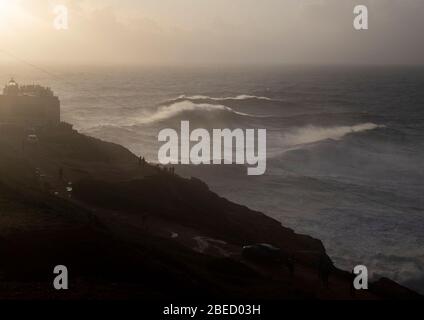 Ein Blick auf Praia do Norte und den berühmten Nazare Leuchtturm, während die Wellen in der untergehenden Sonne zusammenbrechen. Stockfoto