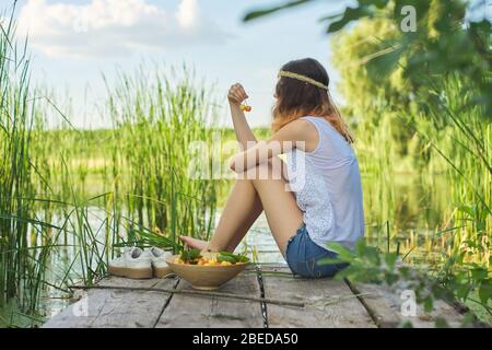 Jugendliches Mädchen sitzt auf Holzsteg mit gelben Kirschen. Schöne junge Frau, die natürliche Früchte isst und den malerischen Sonnenuntergang am See genießt. Schönheit, er Stockfoto