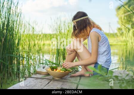 Jugendliches Mädchen sitzt auf Holzsteg mit gelben Kirschen. Schöne junge Frau, die natürliche Früchte isst und den malerischen Sonnenuntergang am See genießt. Schönheit, er Stockfoto