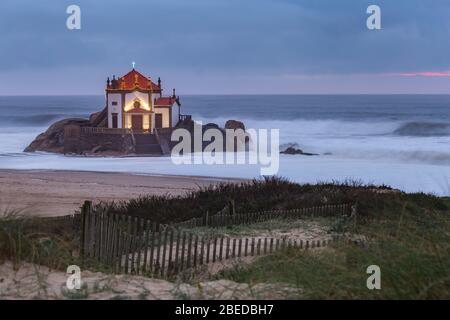 Schöne Kirche am Strand. Capela Senhor da Pedra, bei Sonnenuntergang, mit Wellen über der Kirche Stockfoto