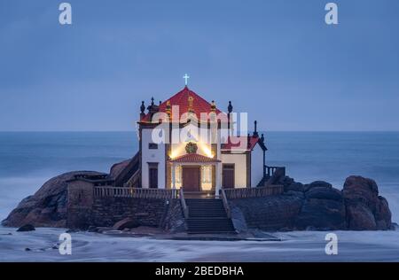 Schöne Kirche am Strand. Capela Senhor da Pedra, bei Sonnenuntergang, mit Wellen über der Kirche Stockfoto