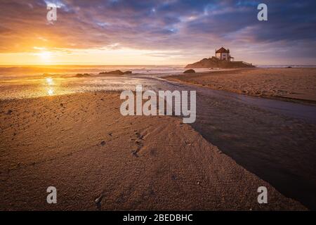 Schöne Kirche am Strand. Capela Senhor da Pedra, bei Sonnenuntergang, mit Wellen über der Kirche Stockfoto