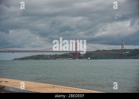 Blick auf die Brücke vom 25. April und das Cristo Rei-Denkmal über dem Tejo unter bewölktem Himmel in Lissabon, Portugal. Stockfoto