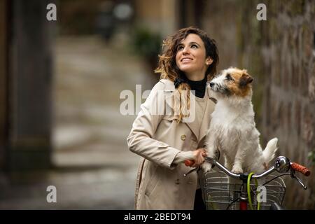 Glückliche junge Frau, die neben ihrem Fahrrad steht und einen Hund im Korb hat Stockfoto