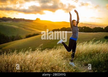 Lächelnde junge Frau springt vor Freude in der Landschaft bei Sonnenuntergang Stockfoto