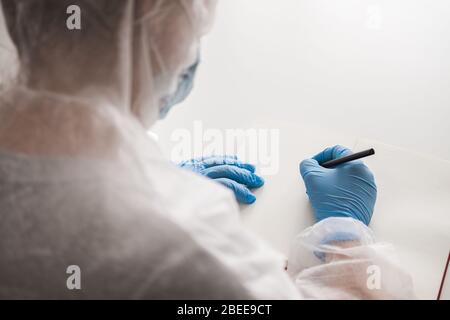 Mädchen im schützenden weißen, lichtdurchlässigen Anzug, blaue Gummihandschuhe, medizinische Maske sitzt zu Hause am Tisch und zeichnet im Skizzenbuch mit Bleistift. Remote-Arbeit von Stockfoto