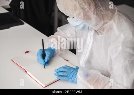 Mädchen im schützenden weißen, lichtdurchlässigen Anzug, blaue Gummihandschuhe, medizinische Maske sitzt zu Hause am Tisch und zeichnet im Skizzenbuch mit Bleistift. Remote-Arbeit von Stockfoto