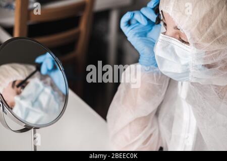 Mädchen im schützenden weißen, lichtdurchlässigen Anzug, blaue Gummihandschuhe, medizinische Maske sitzt zu Hause und malt Wimpern mit Mascara am Spiegel während Stockfoto