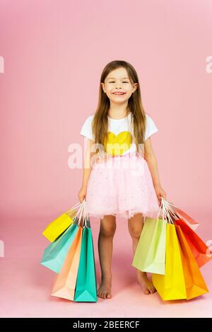 Mädchen mit vielen Einkaufstaschen auf pastellpinkfarbenem Backgound. Schöne süße Momente der kleinen Prinzessin, ziemlich freundliches Kind, das Spaß an der Kamera hat. Stockfoto