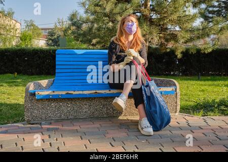 Frau mit medizinischen Maske und Handschuhe sitzt auf einer Bank und macht eine kurze Pause, genießt ein paar Sonnenstrahlen, auf dem Weg nach Hause aus dem Kauf von wichtigen Produkten duri Stockfoto