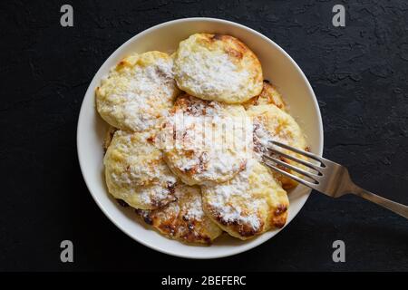 Süße Quark Pfannkuchen auf einem weißen Teller mit Puderzucker bestreut mit einer Gabel auf einem dunklen Hintergrund. Einfach zu hausgemachtes Frühstück zubereiten. Stockfoto