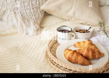 Nahaufnahme eines Tabletts mit Kaffeetasse, Croissants, Müsli und weißen Blumen auf einer Strickdecke im gemütlichen, skandalösen Boho-Schlafzimmer. Bed Breakfast, langsam leben, jom Stockfoto