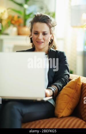 Überrascht stilvolle Frau in weißer Bluse und schwarze Jacke im modernen Wohnzimmer in sonnigen Tag Text auf einem Laptop lesen, während auf der Couch sitzen. Stockfoto