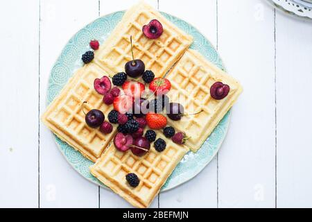 Belgische Waffeln mit Erdbeere, Kirsche, Heidelbeere auf blauem Teller. Weißer Hintergrund. Draufsicht. Stockfoto