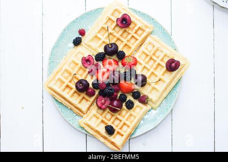 Belgische Waffeln mit Erdbeere, Kirsche, Heidelbeere auf blauem Teller. Weißer Hintergrund. Draufsicht. Stockfoto