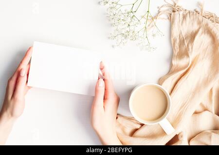 Weibliche Hände halten leere Papierkarte Mockup über weißem Schreibtisch mit Karo und Tasse Kaffee. Hygge, Herbst gemütliche Stimmung, Komfort-Konzept. Flach liegend, Top vie Stockfoto