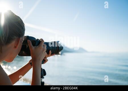 Professionelle Fotografin, die mit ihrer Kamera auf einem Stativ das schöne Morgenmeer fotografiert, wobei aufgehende Sonnenstrahlen über ihrem Kopf scheinen. Stockfoto