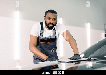 Auto Detaillierung und Reinigung Konzept. Gut aussehende afrikanische männliche Autowascharbeiter, tragen graue Overalls und Handschuhe, wischt und poliert die Auto-Haube mit Stockfoto