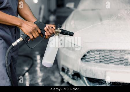 Manuelles Waschkonzept für Autos. Zugeschnittenes Bild von Händen eines jungen dunkelhäutigen Mannes Arbeiter Reinigung modernen weißen Autos mit Schaum und Druck Wasser im Dienst Stockfoto