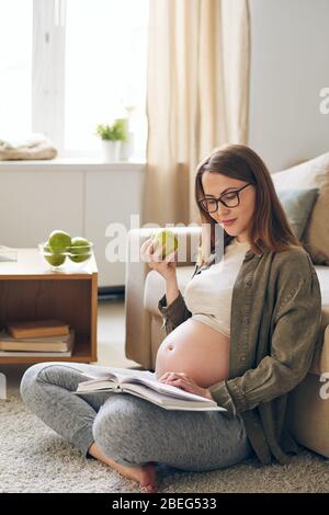 Inhalt Junge schwangere Frau in Brillen sitzt mit gekreuzten Beinen auf dem Teppich und essen grünen Apfel beim Lesen Buch zu Hause Stockfoto