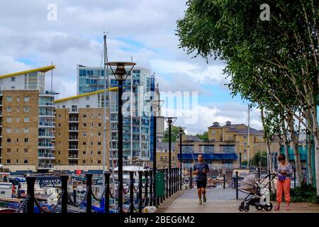 Mann joggen & Dame mit Baby Buggy schaut auf ihr Telefon in Limehouse Basin Marina mit Gebäuden & festverankerten Booten im Hintergrund. Tower Hamlets, London. Stockfoto