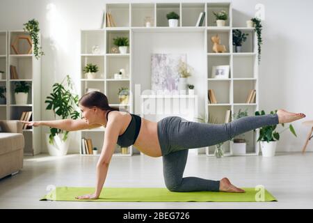 Energetische junge schwangere Frau in Sportbekleidung üben Balancing Tisch Pose auf Yoga-Matte im Wohnzimmer Stockfoto