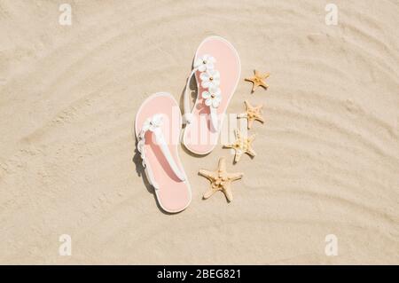 Draufsicht auf weiße Frau Flip Flops mit Blumen und Seesterne auf goldenen Sandstrand in der Nähe des Meeres. Sommerurlaub und Entspannungskonzept. Kopierbereich für Stockfoto