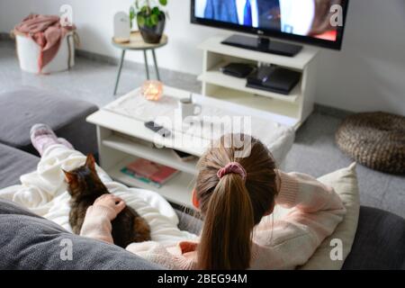 Häusliches Leben mit Haustier. Eine junge Frau sitzt auf der Couch mit ihrer Katze auf ihrem Schoß im Wohnzimmer. Sie schaut fern, während sie ihre Katze streichelte. Stockfoto