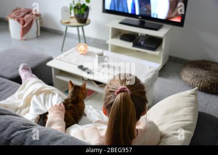 Häusliches Leben mit Haustier. Eine junge Frau sitzt auf der Couch mit ihrer Katze auf ihrem Schoß im Wohnzimmer. Sie schaut fern, während sie ihre Katze streichelte. Stockfoto