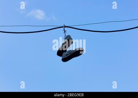 Ein Paar alte Turnschuhe hängen über der Straße von einem elektrischen Kabel, vor einem sauberen blauen Himmel. Stockfoto