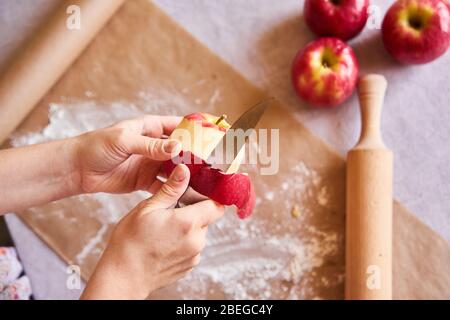 Äpfel, Nudelholz, Mehl, Pergamentpapier auf dem Tisch. Frauenhände schälen einen Apfel. Vorbereitung zum Kochen von Gebäck Stockfoto
