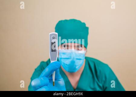 Doktor in grünem Anzug und blauen Maskenhandschuhen mit Thermometer in der Hand auf gelbem Hintergrund. Selektiver Fokus. Stockfoto