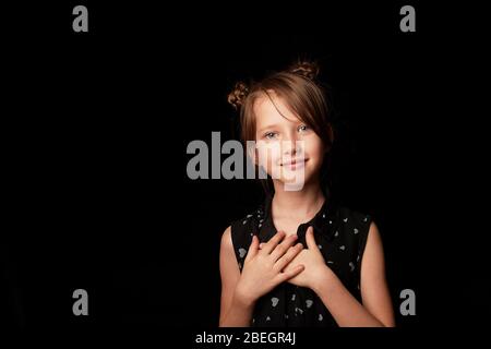 Ein Mädchen von 6 Jahren auf schwarzem Hintergrund. Die Hände sind an der Brust befestigt, ein Zeichen der Annahme und Dankbarkeit. Stockfoto