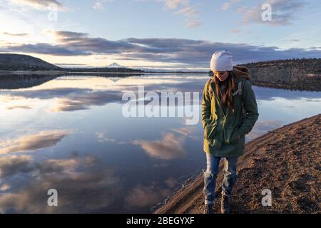 Junge Frau, die bei Sonnenuntergang am See entlang geht, mit Berg im Hintergrund Stockfoto