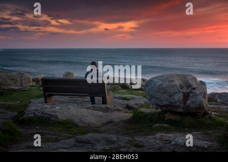 Mann, der einen atemberaubenden Sonnenuntergang auf einer Bank an der Küste beobachtet Stockfoto